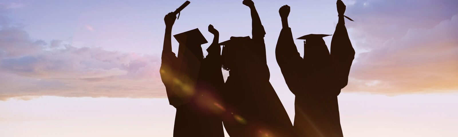 group of graduates in silhouettes with their grad caps on and their hands and diplomas raised